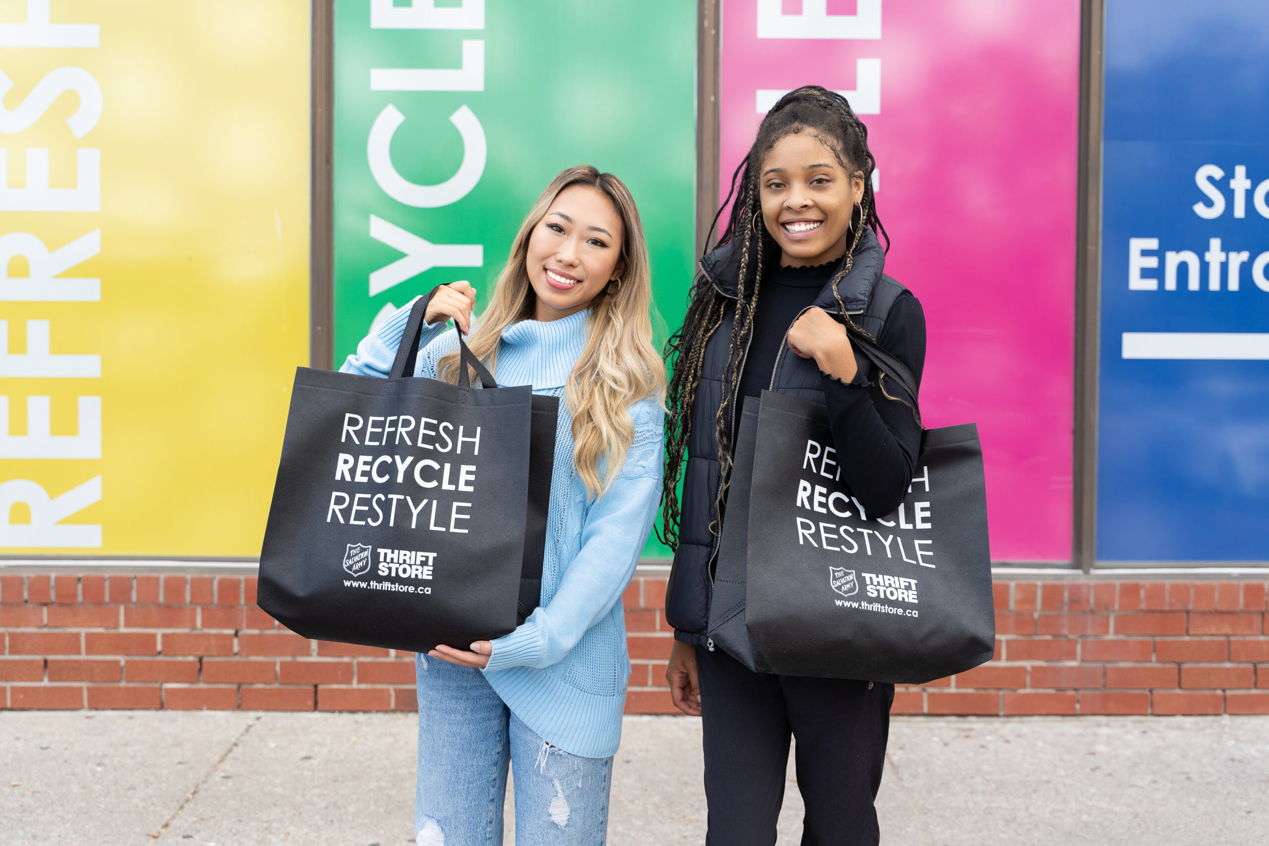 Two females standing outside the Thrift Store holding full reusable bags