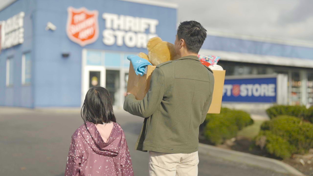 Father holding a box of donations walks toward the Thrift Store with his young daughter