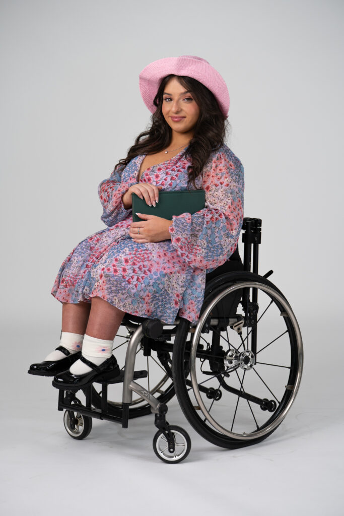 Soft girl outfit model in wheelchair wearing pink and purple floral dress, pink sun hat and holding a book