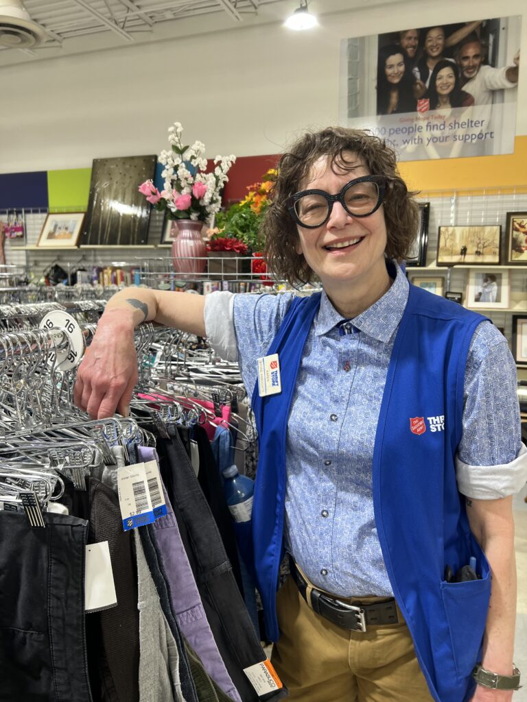 Karen smiling with her arm on a rack of secondhand clothing Salvation Army Thrift Store