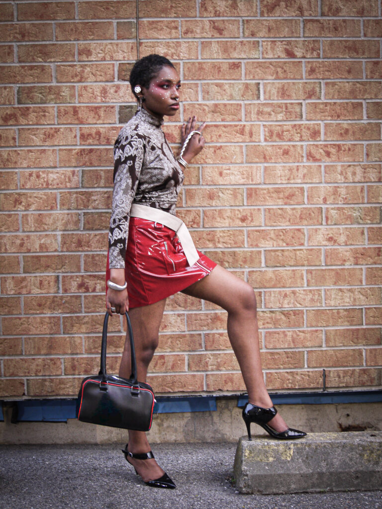 Our collaborator Kendra wearing a mini red skirt and grey and black blouse in front of a brick wall knee bent